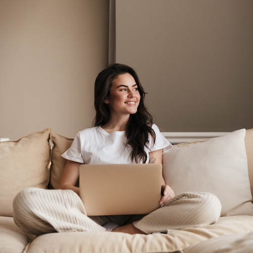 Beautiful smiling young brunette woman relaxing on a couch at home, using laptop computer